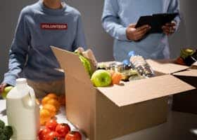 People packing a cardboard box with essentials like fruits, vegetables, and bottled water for charity.
