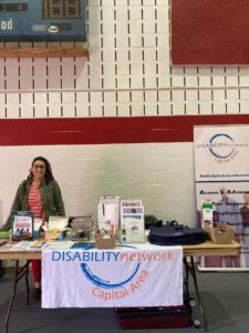 A young white woman with brown hair stands behind a table of DNCAP literature in a gymnasium.