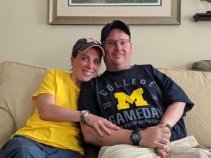 David Pons and his Wife. A white man wearing a blue shirt and a woman wearing a yellow shirt sitting together on a cream colored couch