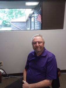 Gill LeMonde- a man with long grey hair pulled back, with a purple polo shirt, seated in his office.