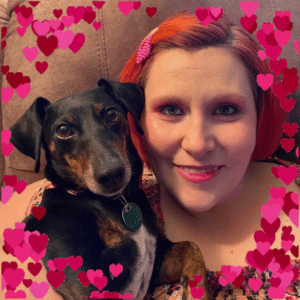 Rachel Burdette-Comer with her dog Frankie. Rachel is a white woman with bright red hair, she is with her dog Frankie and they are seated and surrounded by hearts.