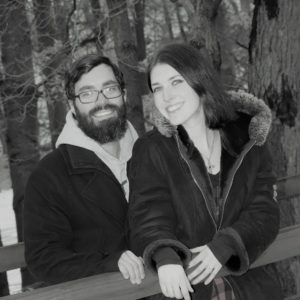 Taylor Phillips and Husband: A black and white photo of a young couple outdoors. She is a young woman with dark hair in a winter coat and he has dark hair and beard with glasses and a winter coat